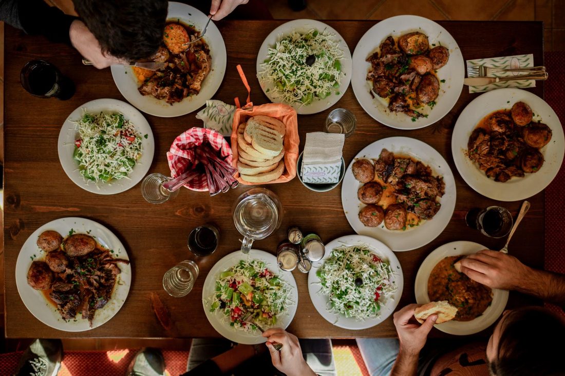 Bovenaanzicht van een houten tafel vol gedeelde borden met vlees, salade en brood terwijl vier mensen samen eten