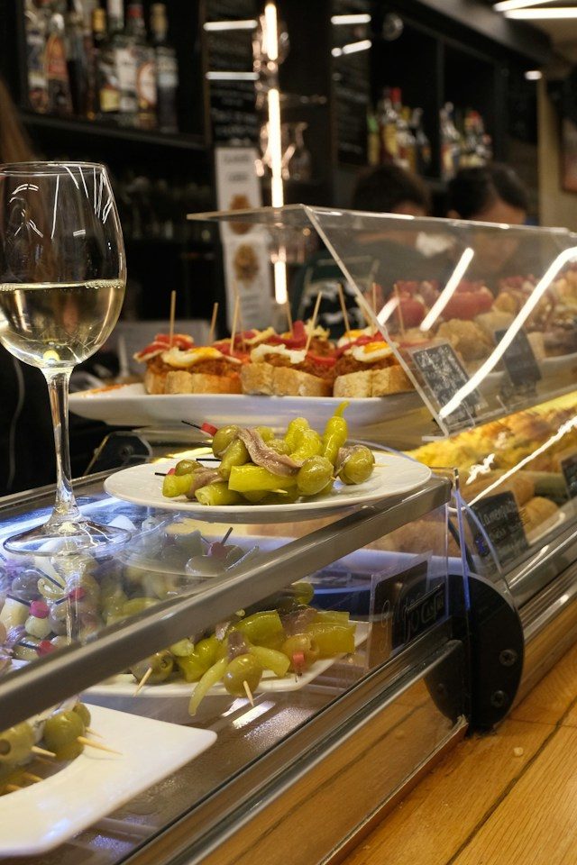 Bar met schalen pintxos en olijvenspiesjes naast een glas witte wijn