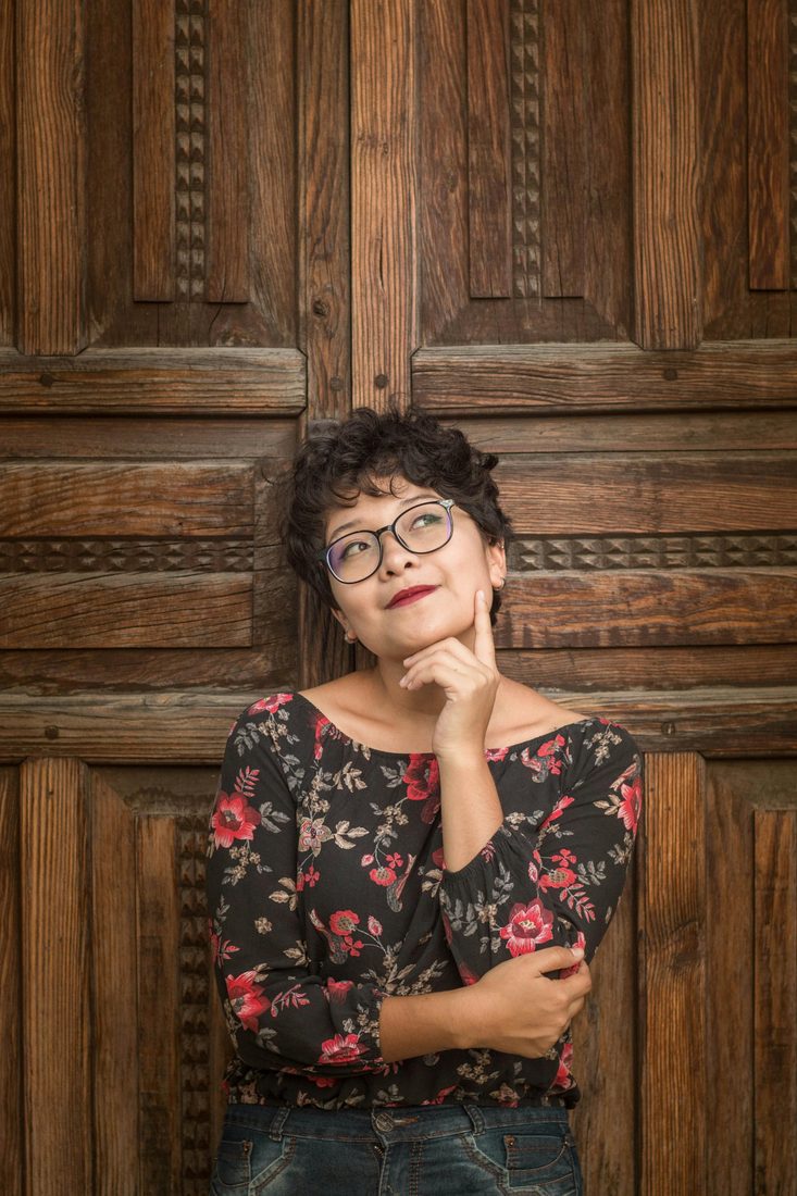 Vrouw met bril leunt tegen een houten deur en kijkt peinzend omhoog