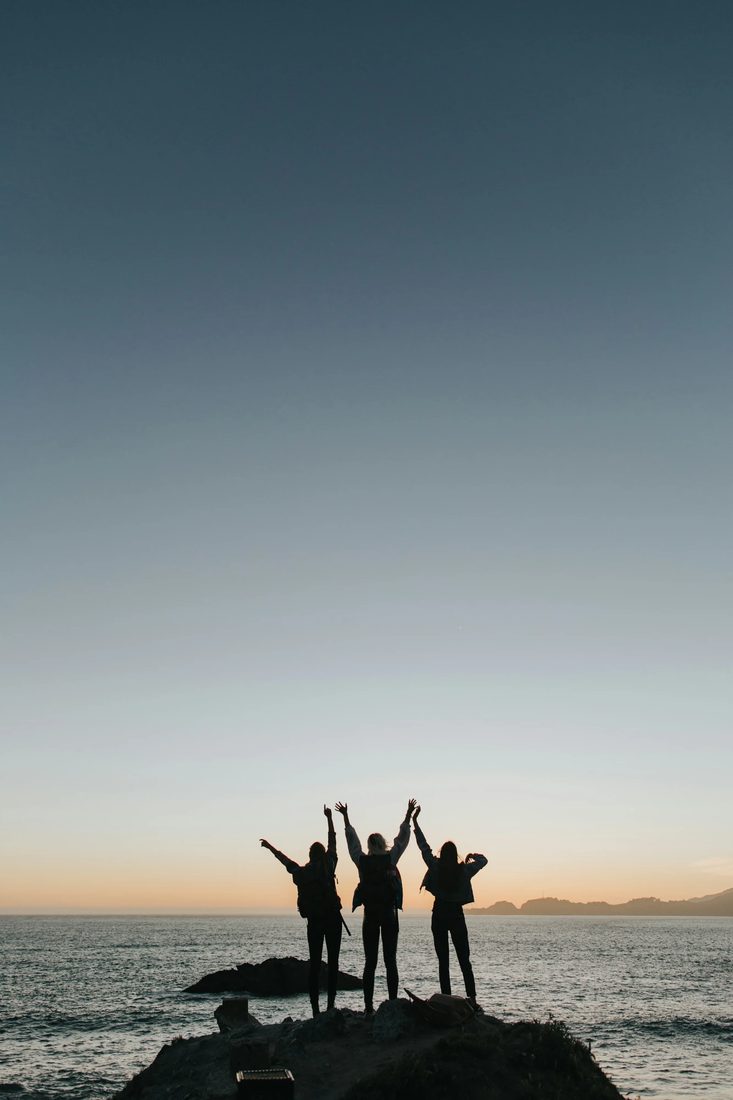 Drie reizigers in silhouet op een rots boven zee, met hun armen omhoog bij zonsondergang