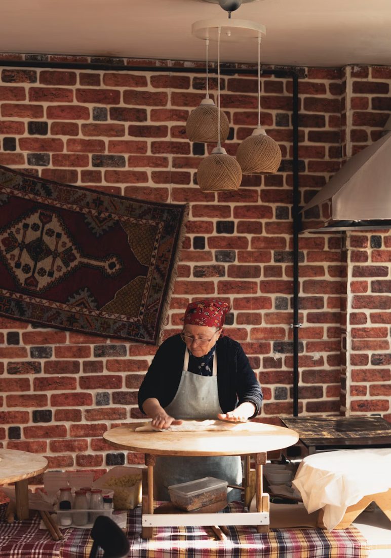 Vrouw met hoofddoek rolt deeg uit op een ronde houten tafel in een keuken met bakstenen muur