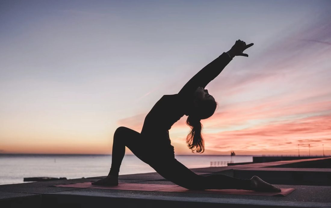 Silhouette of a woman in a yoga pose on a mat at sunset by the water