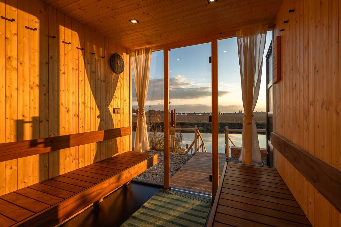 A wooden sauna with open glass doors looking out over the water at sunset