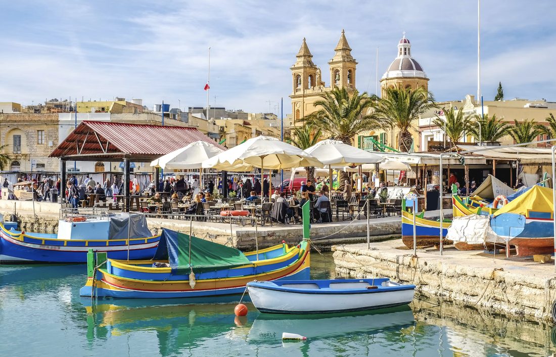 Felgekleurde houten vissersboten in de haven van Marsaxlokk met terrassen en een kerk op de achtergrond